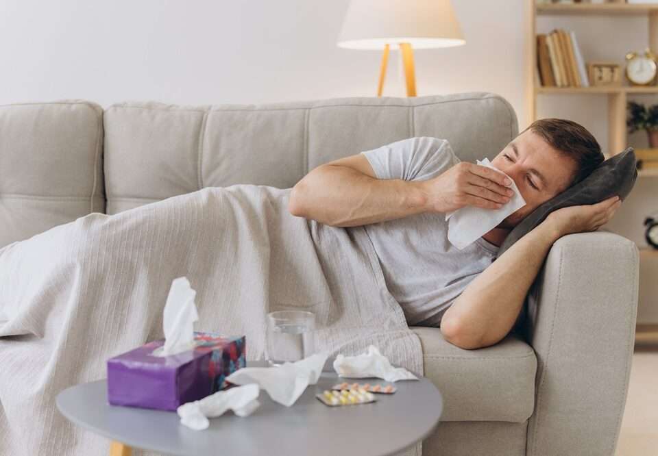 Man lying on a lounge blowing his nose and a table in front with tissues on it