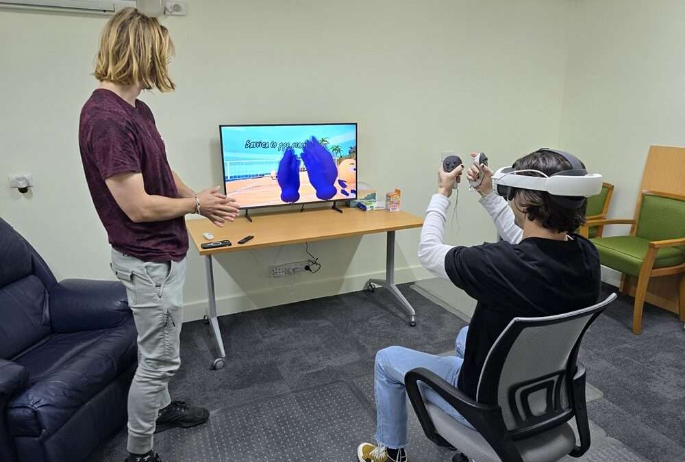 Nathan guiding Cameron through a VR therapy session. Nathan is standing and Cameron is sitting wearing a VR headset in front of a screen