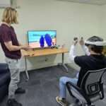 Nathan guiding Cameron through a VR therapy session. Nathan is standing and Cameron is sitting wearing a VR headset in front of a screen