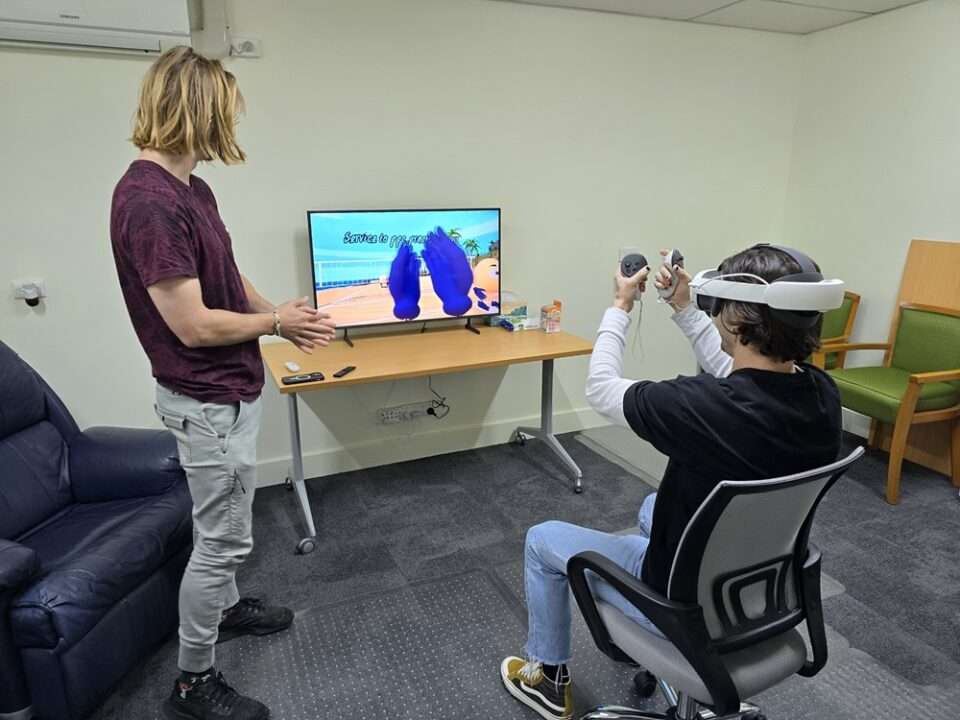 Nathan guiding Cameron through a VR therapy session. Nathan is standing and Cameron is sitting wearing a VR headset in front of a screen